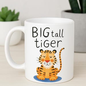 May include: White ceramic mug featuring a cartoon tiger illustration with the text "BIG tall tiger". The tiger is orange with black stripes, sitting on a blue circle. The mug has a curved handle.