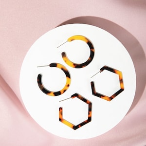May include: Three pairs of earrings on a white background. Two pairs are round hoops with a tortoiseshell pattern. One pair is hexagonal with a tortoiseshell pattern.