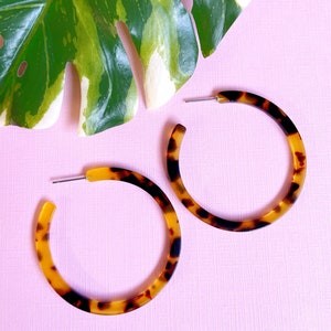 May include: A pair of large, brown and yellow hoop earrings with a tortoiseshell pattern. The earrings are on a pink background.