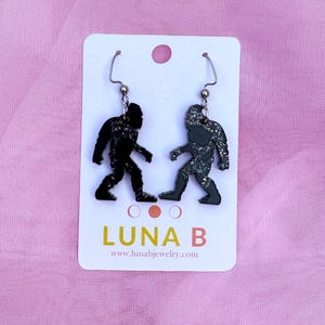 May include: A pair of black glitter Bigfoot earrings with silver hooks. The earrings are on a white card with the text "LUNA B" and "www.lunabjewelry.com".