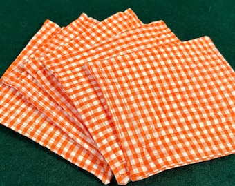May include: A stack of four square fabric coasters featuring a bright orange and white gingham checkered pattern. Each coaster has stitched edges and is arranged in a slightly fanned, overlapping formation on a dark green surface.