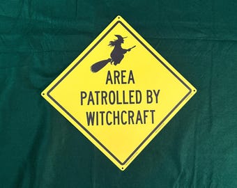 May include: A diamond-shaped yellow metal sign with a black border and a black silhouette of a witch flying on a broomstick. The text in black capital letters reads AREA PATROLLED BY WITCHCRAFT. The sign has small mounting holes at the corners.
