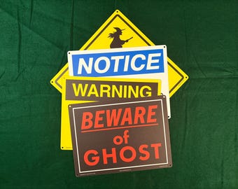 May include: A collection of four rectangular and diamond-shaped metal signs. The top sign is yellow with a black witch silhouette. Below it, a white sign reads NOTICE in blue. A yellow sign reads WARNING in black, and a black sign reads BEWARE of GHOST in red.