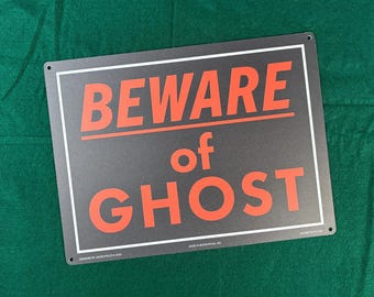 May include: A rectangular black metal sign with a white border and the text BEWARE of GHOST printed in bold orange capital letters. The sign features small circular holes in each corner for mounting and includes small white text at the bottom.