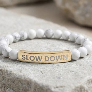 May include: A beaded bracelet featuring white and gray marbled beads. A gold-colored bar in the center is engraved with the words "SLOW DOWN". The bracelet is resting on a gray stone surface.