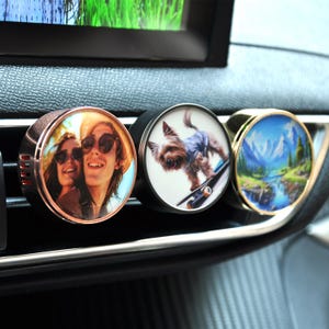 May include: Three car air fresheners with different designs. One has a photo of two people, another features a dog on a skateboard, and the third displays a landscape with mountains and a river. The fresheners are round and metallic.