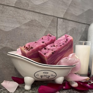 May include: Two pink and white swirl soap bars with red flecks in a white porcelain bathtub-shaped dish with the words "Le Bain" on the front. The dish is surrounded by pink rose petals.