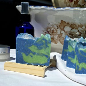 May include: Three bars of handmade soap with a blue and green swirl pattern. The soap is sitting on a wooden soap dish. There is a blue glass bottle with a spray nozzle in the background.