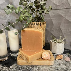 May include: Two bars of handmade soap with a light orange hue rest on a small wooden soap dish. A seashell and a small pink crystal are next to the dish. In the background are two candles in glass holders, a woven basket with greenery, and a small potted plant.