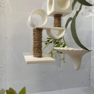 May include: A white plastic hamster cage with a multi-level play structure. The structure includes a platform, a tube, a small house, and a hanging dish. The cage is decorated with a small green plant and a piece of brown rope.