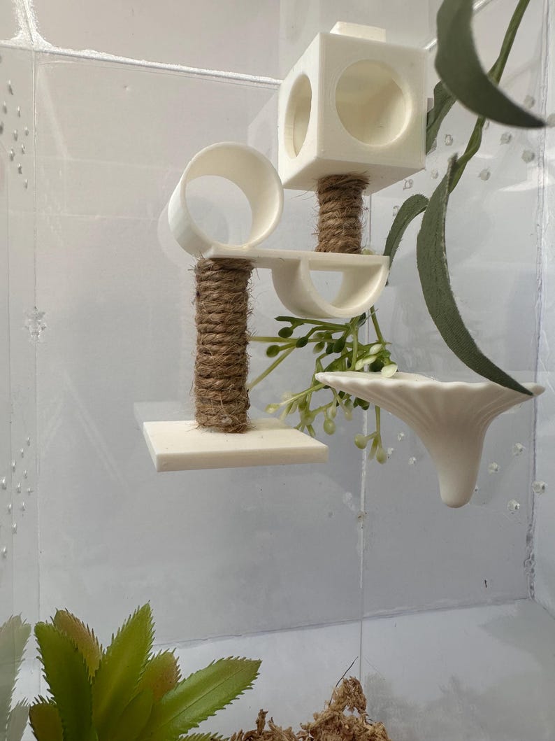 May include: A white plastic hamster cage with a multi-level play structure. The structure includes a platform, a tube, a small house, and a hanging dish. The cage is decorated with a small green plant and a piece of brown rope.