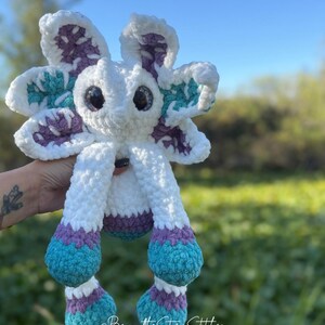 May include: A handmade crochet plush toy in the shape of a whimsical creature. The toy features a white body with large, expressive eyes. The flower-like head and feet are accented with teal, purple, and white. The text "@BeyondtheStarsStitches" is visible.
