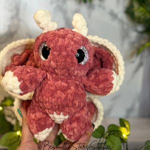 May include: A crocheted plush toy in the shape of a pink dragon with white horns and white accents. The dragon has large black eyes with white pupils. The dragon is sitting in a crocheted nest.