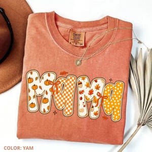 May include: A yam-colored short-sleeve t-shirt featuring the word MAMA in large, decorative letters. The letters are filled with patterns of pumpkins, autumn leaves, orange and white checkers, and polka dots, accented with orange bows.