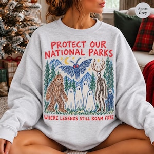 May include: A sport grey crewneck sweatshirt featuring a crayon-style graphic with the red text "PROTECT OUR NATIONAL PARKS" and "WHERE LEGENDS STILL ROAM FREE". The illustration depicts a mothman, bigfoot, ghosts, and a deer-like creature.