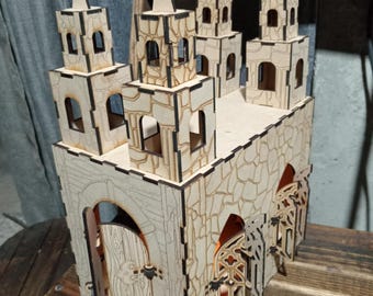 May include: A laser-cut wooden model of a church with multiple towers, crosses, and arched windows. The model features detailed stonework and open doorways. The church is made of light-colored wood and sits on a wooden surface.