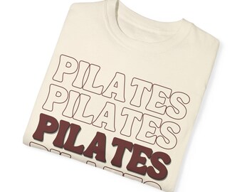 May include: A cream colored t-shirt with the word "PILATES" repeated four times in a stacked, retro font. The text is brown with a white outline.