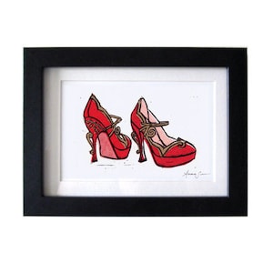 May include: A framed print of a red and gold illustration of two high-heeled shoes. The shoes have a strap across the top and a decorative design on the heel. The print is signed "Anna Jean".