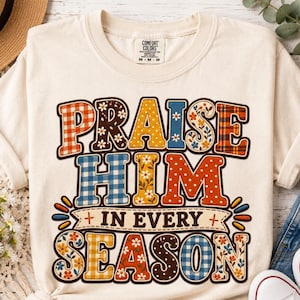 May include: A cream-colored short-sleeve t-shirt featuring a multi-colored graphic print with the text "PRAISE HIM IN EVERY SEASON". The letters are filled with various patterns including plaid, polka dots, and floral designs in red, blue, and yellow.