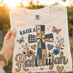 Puede incluir: Una camiseta color crema con un gráfico de una cruz estilo patchwork, mariposas, flores y corazones. El texto "RAISE A Hallelujah" aparece en letras azules y marrones. El diseño incluye patrones de cuadros vichy azules y blancos y flores.
