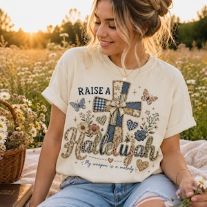May include: A cream-colored short-sleeve t-shirt featuring a graphic of a cross made of blue and tan patterned fabric, surrounded by flowers, butterflies, and hearts. The text reads "RAISE A Hallelujah" and "My weapon is a melody" in dark blue.