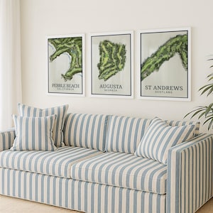 May include: Three framed wall art prints featuring aerial views of golf courses labeled Pebble Beach California, Augusta Georgia, and St Andrews Scotland, displayed above a sofa upholstered in a blue and white vertical striped fabric.