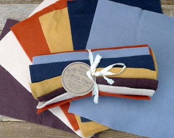 May include: A collection of wool felt sheets in various neutral colors, including shades of blue, orange, and cream. The felt is bundled and tied with a white ribbon, with a tag that reads "Wool Felt". The sheets measure 9 x 12 inches.