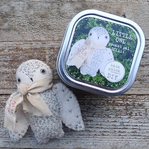 May include: A white felt owl DIY kit in a silver tin. The kit includes felt pieces, embroidery floss, and instructions to create a small owl with a white scarf. The tin has a label that reads "Little Owl Pocket Pal DIY Kit" and "kata golda handmade DIY".