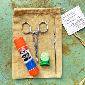 May include: A small tan fabric pouch with a drawstring closure contains a set of sewing tools. The tools include a pair of hemostat forceps, a green thimble, a crochet hook, and a glue stick. The pouch has a tag attached that reads "imperfect patchwork & little stitchings a kit for mending, decorating, and creating extra tools hemostat forceps for pulling a needle through thick fabric thimble [ML] crochet hook glue stick"