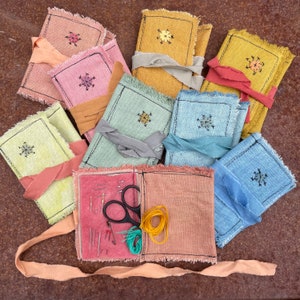 May include: A collection of colorful, handmade sewing kits. Each kit features a linen cover in shades of pink, yellow, blue, and green, with a cross-stitch design. The kits are tied with matching ribbons and contain needles, scissors, and thread.