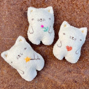 May include: Three small, white, plush cat-shaped toys with embroidered details. One cat holds a pink flower, one holds an orange heart, and one holds a yellow star. Each cat has gold-colored fabric accents on the ears and simple stitched facial features.