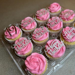 May include: A clear plastic container filled with pink frosted cupcakes decorated with silver glitter lettering that says "Happy Birthday". One cupcake has a silver glitter number "19" on top.