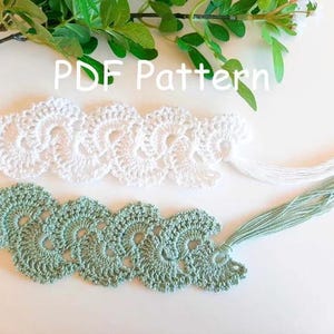 以下が含まれることがあります： 2つのクロシェのブックマーク。1つは白、もう1つは緑で、両端にタッセルが付いています。ブックマークは繊細なレース模様でできています。画像には「PDF Pattern」というテキストが表示されています。