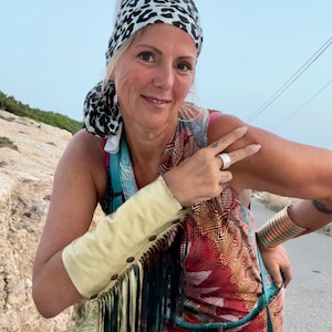 Könnte beinhalten: Eine Frau trägt ein Kopftuch mit Leopardenmuster, einen farbenfrohen, gemusterten Jumpsuit, eine Lederarmbinde mit Fransen und eine türkisfarbene Tasche. Sie macht ein Peace-Zeichen mit ihrer Hand und trägt einen Ring an ihrem Finger.