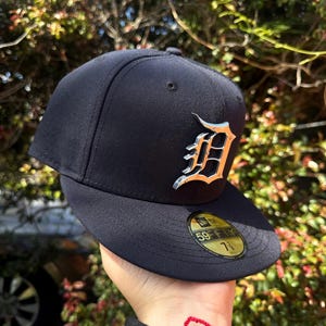 Puede incluir: Una gorra de béisbol negra con un logotipo D de metal plateado. La gorra tiene una visera negra y una etiqueta 59FIFTY dorada.