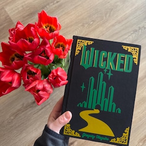 Puede incluir: Un libro de tapa dura negro con el título "WICKED" en letras verdes y doradas. La portada del libro presenta una silueta verde de Ciudad Esmeralda y el Camino de Baldosas Amarillas. El libro es de Gregory Maguire.