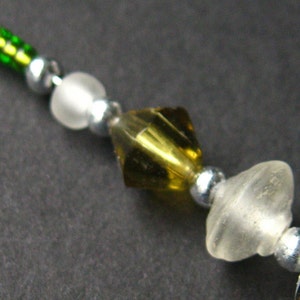 May include: A close-up of a beaded necklace with green, white, and gold beads. The beads are strung on a silver wire.