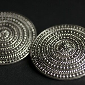 May include: A pair of silver-toned earrings with a circular design featuring concentric circles and a central flower motif.