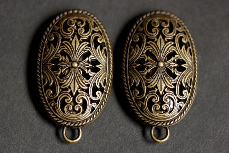 May include: Two oval-shaped, antique gold-toned metal embellishments with intricate floral designs. Each embellishment has a loop on the bottom.