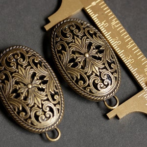May include: Two antique gold-toned metal oval-shaped decorative embellishments with intricate floral designs. The embellishments are approximately 2.5 inches long and 1.5 inches wide.