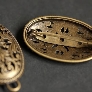 May include: Two antique gold-toned oval brooches with intricate filigree designs. One brooch has a pin closure and the other has a loop closure.