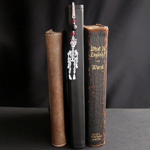May include: A silver skeleton bookmark with red and black beads hangs from a book titled "What Is English?" by Ward.