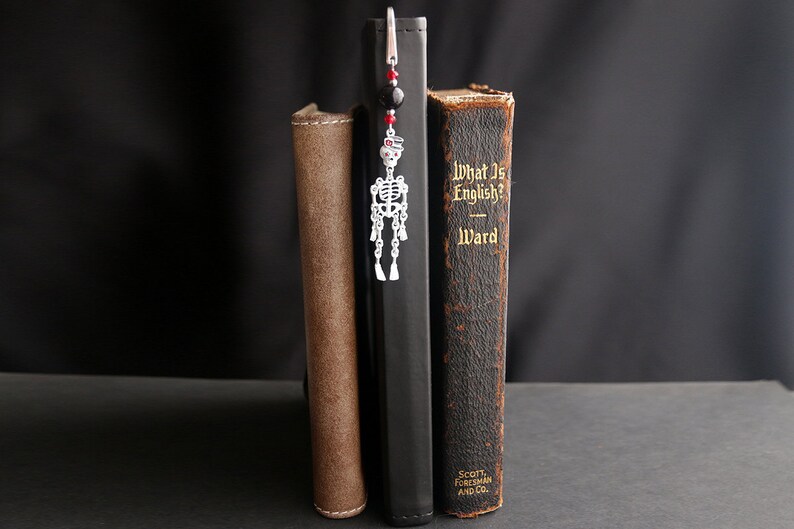 May include: A silver skeleton bookmark with red and black beads hangs from a book titled "What Is English?" by Ward.