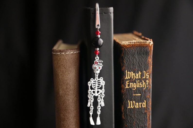 May include: A silver skeleton bookmark with red and black beads hanging from a silver clip. The bookmark is hanging from the spine of a book titled "What Is English? Ward".