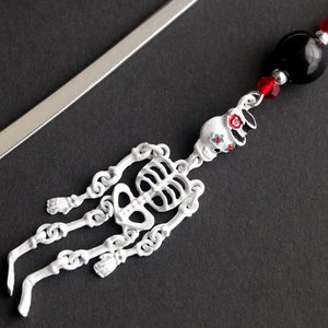 May include: A silver bookmark with a white skeleton charm and black and red beads. The skeleton charm has a floral design on the skull.