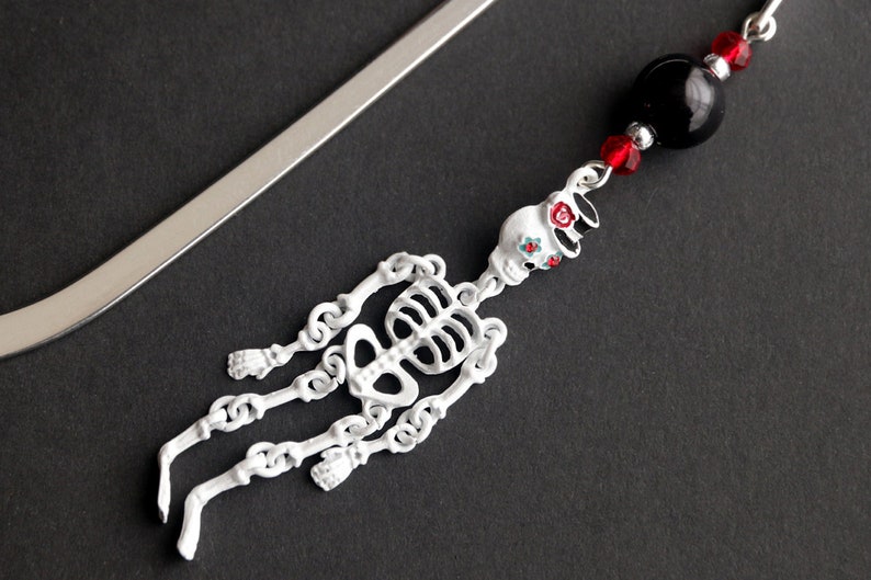 May include: A silver bookmark with a white skeleton charm and black and red beads. The skeleton charm has a floral design on the skull.