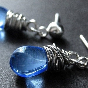 Può includere: Un paio di orecchini d'argento con perle di vetro blu a forma di lacrima avvolte in filo d'argento. Gli orecchini hanno un semplice chiusura a perno.