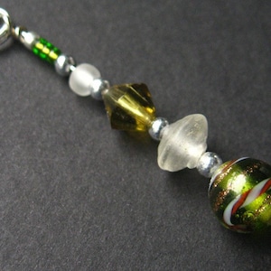 May include: A green, white, and gold glass bead charm with a silver ring attached. The charm is made of several different beads, including a clear glass bead, a gold and green glass bead, and a green and white glass bead.