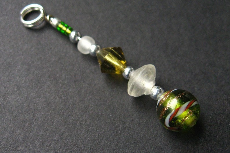 May include: A green, white, and gold glass bead charm with a silver ring attached. The charm is made of several different beads, including a clear glass bead, a gold and green glass bead, and a green and white glass bead.