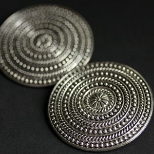 May include: Two silver circular earrings with intricate embossed designs. The earrings feature concentric circles of raised dots and a central flower-like motif.